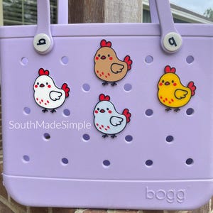 May include: A lavender tote bag with four cartoon chicken charms. The bag has two handles and the word "bogg" printed on the bottom. The text "SouthMadeSimple" is printed on the bag.