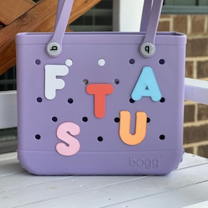 May include: A purple tote bag with the word "bogg" printed on the bottom. The bag has a variety of colorful letters attached to it, spelling out "FTASU".