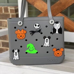 May include: A gray tote bag with the word "bogg" printed on the bottom. The bag has various Halloween-themed characters attached to it, including Mickey Mouse, a ghost, a witch, and Jack Skellington.