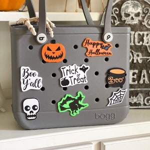 May include: A gray tote bag with a variety of Halloween-themed stickers, including a pumpkin, a witch, a ghost, a spiderweb, and a cauldron. The bag has the word "BOGG" printed on the bottom.