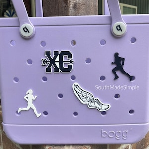 May include: A lavender-colored tote bag with a white handle and a white 'bogg' logo. The bag has a variety of white and black stickers, including a running figure, a winged shoe, and a cross country logo. The text 'SouthMadeSimple' is also visible on the bag.