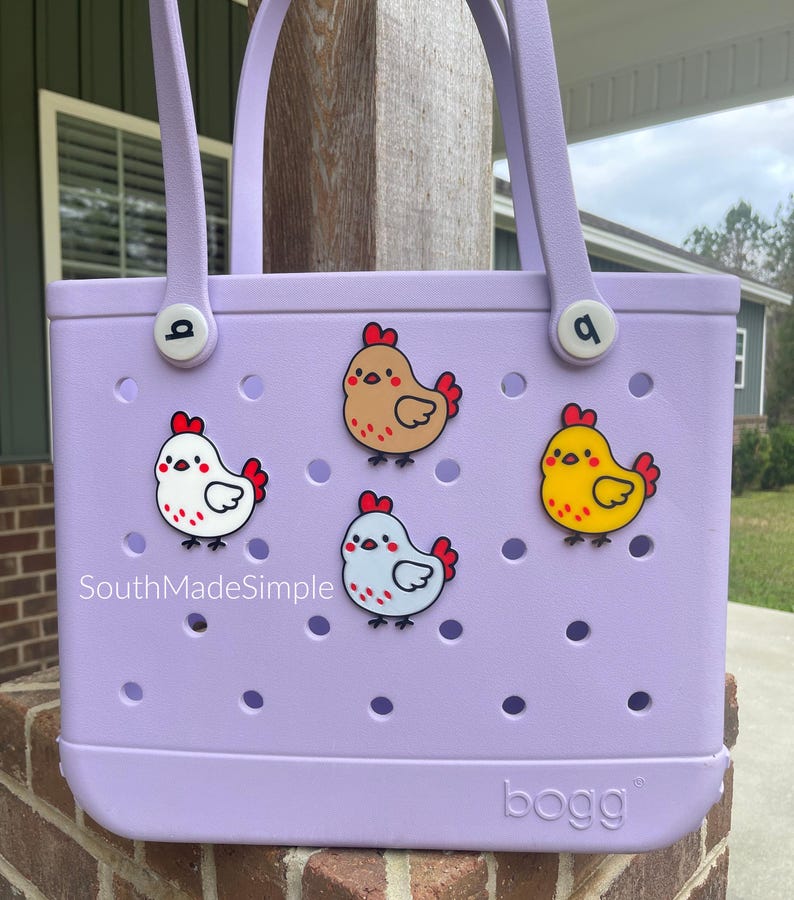 May include: A lavender tote bag with four cartoon chicken charms. The bag has two handles and the word "bogg" printed on the bottom. The text "SouthMadeSimple" is printed on the bag.