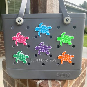 Puede incluir: Bolso tote gris decorado con abalorios con forma de tortuga de colores. Los abalorios son en tonos rosa, azul, verde, morado y naranja. El bolso tiene un asa y la palabra "bogg" es visible en la parte inferior. El texto "SouthMadeSimple" tambi&eacute;n es visible.