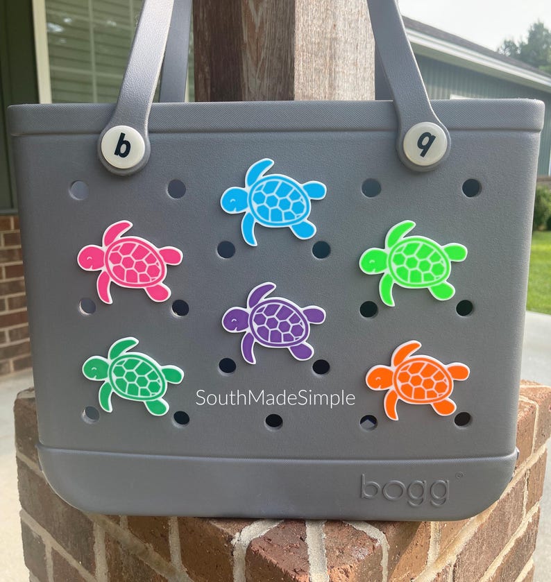 Puede incluir: Bolso tote gris decorado con abalorios con forma de tortuga de colores. Los abalorios son en tonos rosa, azul, verde, morado y naranja. El bolso tiene un asa y la palabra "bogg" es visible en la parte inferior. El texto "SouthMadeSimple" tambi&eacute;n es visible.