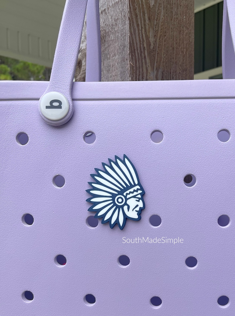 May include: A lavender tote bag with a white and blue Native American headdress charm. The bag has a white button with a circle design and a white text label that reads "SouthMadeSimple".