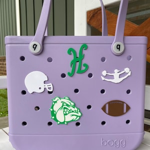 May include: A lavender-coloured tote bag with a "bogg" logo. The bag has a variety of green, white, and brown patches, including a bulldog, a football, a cheerleader, and a letter "H".
