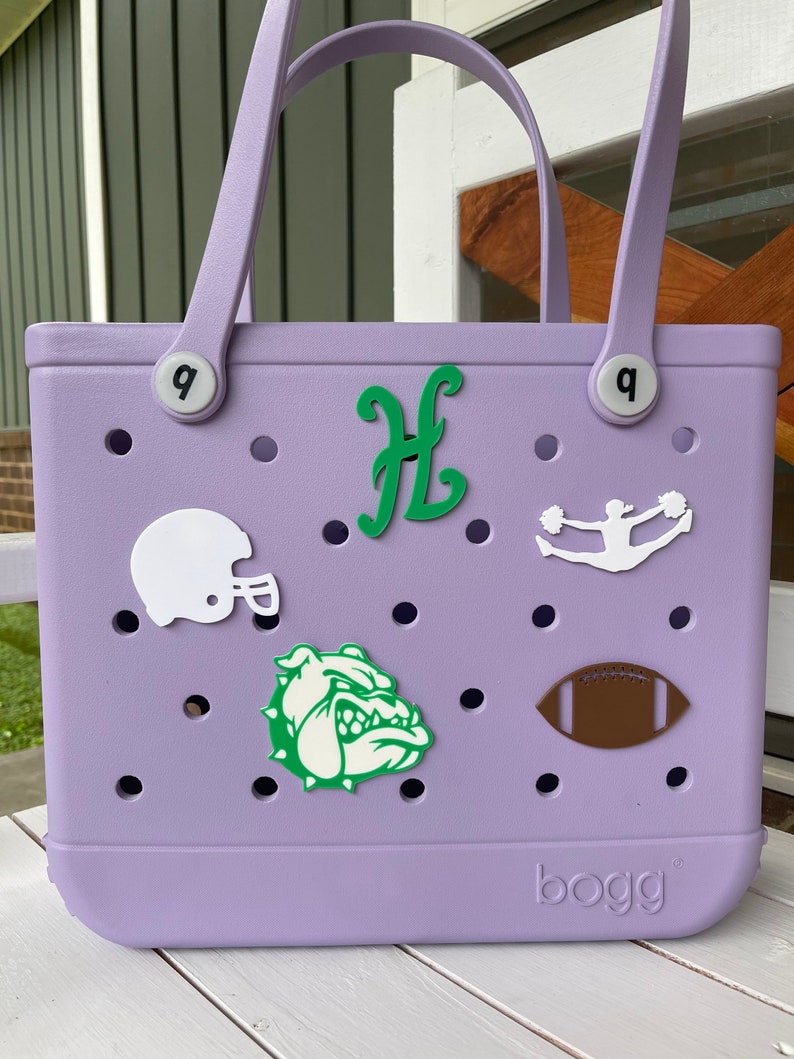 May include: A lavender-coloured tote bag with a "bogg" logo. The bag has a variety of green, white, and brown patches, including a bulldog, a football, a cheerleader, and a letter "H".