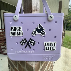 May include: A light purple tote bag with white accents and a handle. The bag has several white stickers with the phrases "RACE MAMA" and "DIRT LIFE", a motorcycle graphic, and crossed checkered flags. The bag is made of a durable material.