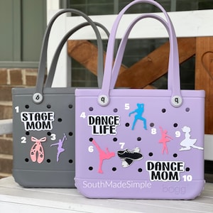 May include: Two purple and gray tote bags with holes for adding charms. The purple bag has the text "Dance Life" and "Dance Mom" on it. The gray bag has the text "Stage Mom" on it. Both bags have various dance-themed charms attached.