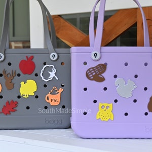 May include: Two gray and purple tote bags with a variety of fall-themed decorations, including a pumpkin, an apple, a turkey, a deer, a cotton boll, a coffee cup, a corn cob, an owl, a squirrel, and an acorn. The bags are both labeled "bogg".