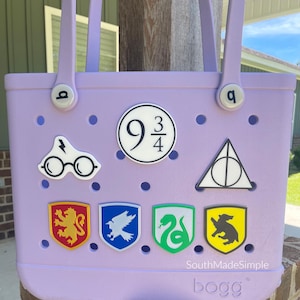 May include: A lavender tote bag with a variety of Harry Potter-themed charms. The charms include the 9 3/4 logo, glasses and lightning bolt, and house crests. The bag has two handles and the brand name "bogg" is visible.