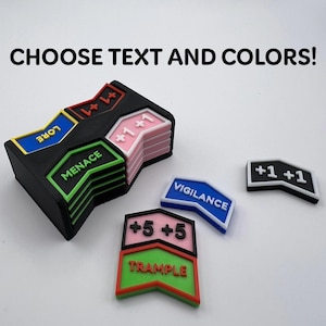 May include: A stack of black plastic game tokens with coloured inserts featuring text like LORE, MENACE, and +1 +1. Loose tokens include VIGILANCE, +5 +5, TRAMPLE, and +1 +1. The text CHOOSE TEXT AND COLOURS! is displayed at the top of the image.