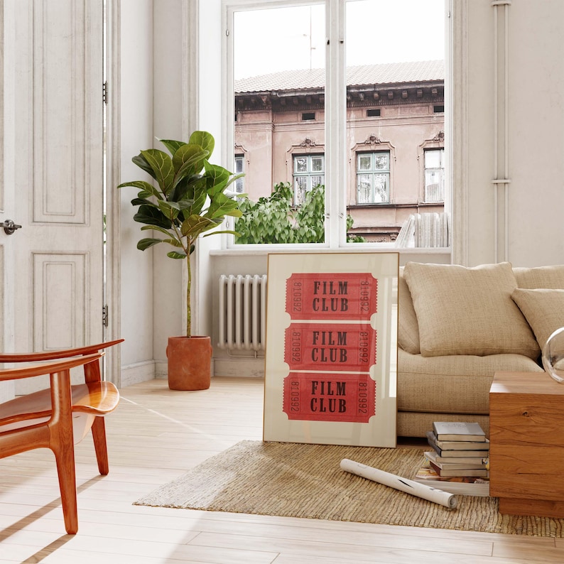 May include: A framed print with the text "FILM CLUB" repeated three times in a red ticket design. The print is hanging on a white wall in a living room with a couch, a chair, and a plant.