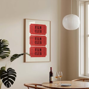 May include: A framed print with three red tickets that say "Film Club" in black text. The tickets are in a wooden frame and are hanging on a beige wall. There is a glass of wine and a bottle of wine on a wooden table in front of the print.