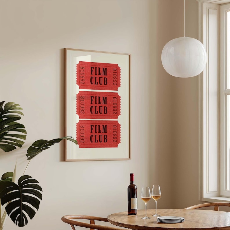 May include: A framed print with three red tickets that say "Film Club" in black text. The tickets are in a wooden frame and are hanging on a beige wall. There is a glass of wine and a bottle of wine on a wooden table in front of the print.