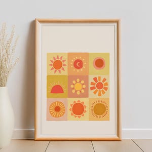 May include: A framed art print with a grid of nine sun illustrations in orange, yellow, and pink. The artwork is in a light wood frame against a neutral wall. A vase with dried flowers is on the left. The frame is approximately 50 cm in height.