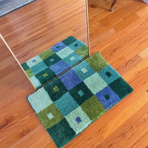 Puede incluir: Una alfombra geométrica con un diseño cuadrado en verde, azul y verde claro. La alfombra está sobre un suelo de madera.