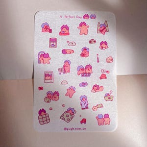 May include: A sticker sheet featuring the text "A Perfect Day" and multiple illustrations of a cartoon character. The illustrations are in pink, purple, and white. The artist's name, purple.times.art, is at the bottom of the sheet.