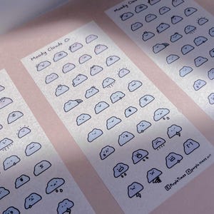 May include: Three rectangular sticker sheets featuring rows of small, light blue cartoon clouds with various facial expressions and weather elements. Each sheet is titled Moody Clouds and includes the text Purple Times at the bottom right.