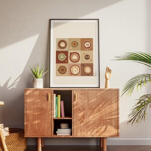 May include: A framed art print with nine coffee-themed illustrations in a grid pattern, in brown, beige, and green tones. It's displayed above a wooden cabinet with gold handles, a small succulent, and a wooden hand sculpture. A potted plant is to the right.