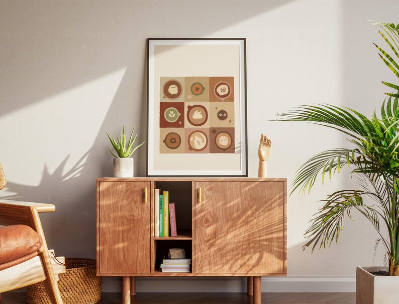 May include: A framed art print with nine coffee-themed illustrations in a grid pattern, in brown, beige, and green tones. It's displayed above a wooden cabinet with gold handles, a small succulent, and a wooden hand sculpture. A potted plant is to the right.
