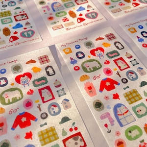 May include: Sheets of decorative stickers with colourful illustrations. The stickers depict a red sweater, a cat, a house, a sheep, and the text "My Favorite Things". The stickers are on a white background.