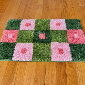 Peut inclure: Un tapis à carreaux rose et vert avec une texture duveteuse. Le tapis est fait d'un matériau doux et moelleux et présente un motif géométrique.