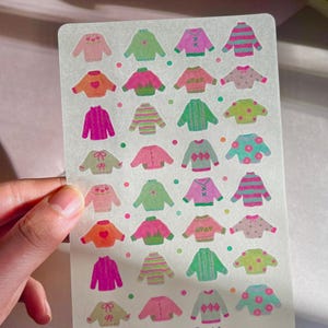 May include: A sticker sheet with illustrations of sweaters in shades of pink, green, and orange. The sweaters have diverse patterns, including stripes, hearts, and argyle. The text "purple.times.art" and "Purple Times" are printed on the sheet.