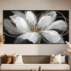May include: A large, framed canvas art piece featuring a white and gray flower with gold accents against a black background. The artwork hangs above a modern sofa with neutral-colored cushions. The room is decorated with natural elements.