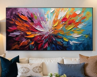 May include: Large abstract painting with a black frame, featuring a burst of vibrant colors including red, orange, yellow, white, and blue. The artwork is displayed above a white sofa with decorative pillows, creating a modern living room aesthetic.