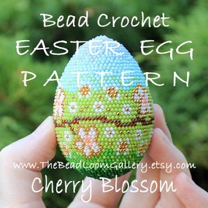 以下が含まれることがあります： 桜の花のデザインが施されたビーズクロシェのイースターエッグのパターン。卵は青、緑、白で、茶色のアクセントが付いています。パターンは「チェリーブロッサム」と呼ばれています。