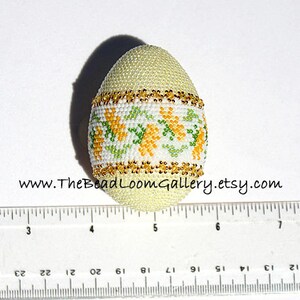 May include: A beaded egg with a pale yellow base, adorned with a band of white, green, and yellow floral patterns. Gold-colored beads trim the top and bottom of the floral band. The website address www.TheBeadLoomGallery.etsy.com is visible.