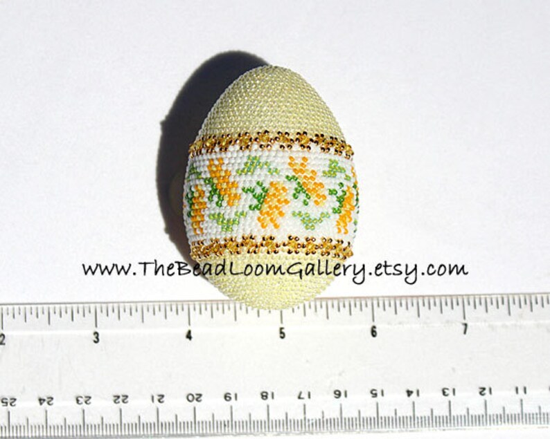 May include: A beaded egg with a pale yellow base, adorned with a band of white, green, and yellow floral patterns. Gold-colored beads trim the top and bottom of the floral band. The website address www.TheBeadLoomGallery.etsy.com is visible.