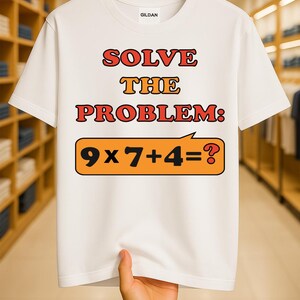 May include: White t-shirt with the text "SOLVE THE PROBLEM: 9 x 7 + 4 = ?" in red and orange lettering. The shirt is held up by a hand, showcasing the design. The shirt is made of a soft material.