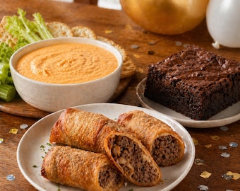 Puede incluir: Una selección de aperitivos en una mesa de madera. Incluye un cuenco con salsa naranja, galletas saladas, apio, un brownie y rollitos de primavera. Un rollito de primavera está cortado por la mitad, revelando el relleno. Globos dorados y blancos en el fondo.