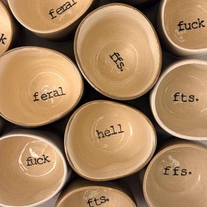 May include: A collection of cream-colored ceramic bowls with irregular, organic shapes. Each bowl features a smooth, glossy interior finish and is stamped with black text at the bottom, including the words feral, fts., fuck, ffs., and hell.