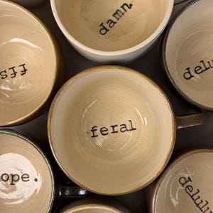 May include: A collection of ceramic mugs with speckled cream interiors and handles. Each mug features a unique word stamped in black ink at the bottom, including the words damn, ffs, feral, nope, delulu, and wtf, all viewed from above.