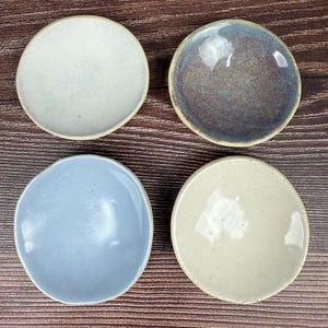 May include: Four small, round ceramic dishes in varying shades of blue, gray, and cream. Each dish has a slightly irregular shape and a raised rim. The dishes are arranged on a dark wood-grain surface.