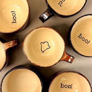 May include: A collection of six ceramic mugs with dark brown glazed exteriors and cream-colored interiors. Four mugs feature the word "boo!" printed in black at the bottom, one mug has a simple black ghost outline, and one is plain inside.