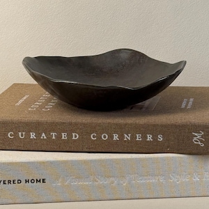 May include: A dark brown, shallow bowl with an irregular, wavy rim. The bowl has a matte, textured surface and sits atop a brown book titled Curated Corners, which is stacked on a light-colored book with a yellow and white patterned cover.