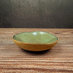 Puede incluir: Un pequeño cuenco de cerámica hecho a mano con forma redondeada. El interior tiene un esmalte verde claro brillante, mientras que el exterior tiene un acabado marrón terroso texturizado. El cuenco está sobre una superficie de madera.