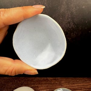 以下が含まれることがあります： 淡い青色の楕円形セラミック皿。暗い背景を背景に、手で皿を持っています。皿は滑らかで光沢のある表面をしています。他の同様の皿が下に表示されています。