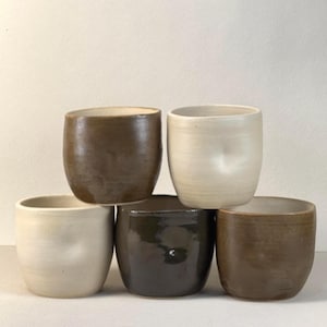 May include: A set of five ceramic cups arranged in a stack. The collection includes two off-white cups with a subtle thumb indentation, two brown cups with a smooth finish, and one dark brown cup with a glossy, reflective surface.