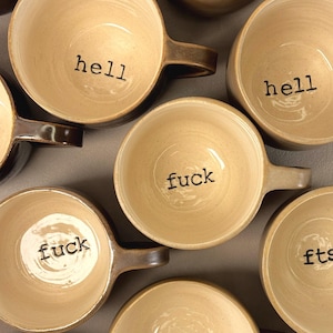 May include: A collection of ceramic mugs with a light beige interior and a darker brown exterior. Each mug features a single word stamped in black ink at the bottom of the interior, including the words hell, fuck, fts, and wtf, all in lowercase.