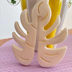 Könnte beinhalten: Ein weißes, 3D-gedrucktes, blattartiges Objekt mit einem detaillierten Design. Das Blatt liegt auf einer violetten, gewebten Oberfläche.