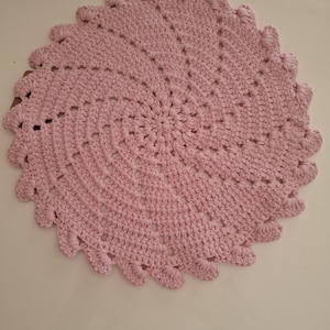 May include: A handmade, light pink crocheted doily. The circular doily features a spiral design with a scalloped edge. The texture of the crochet stitches is visible, creating a soft, tactile appearance. This decorative item is suitable for home decor.