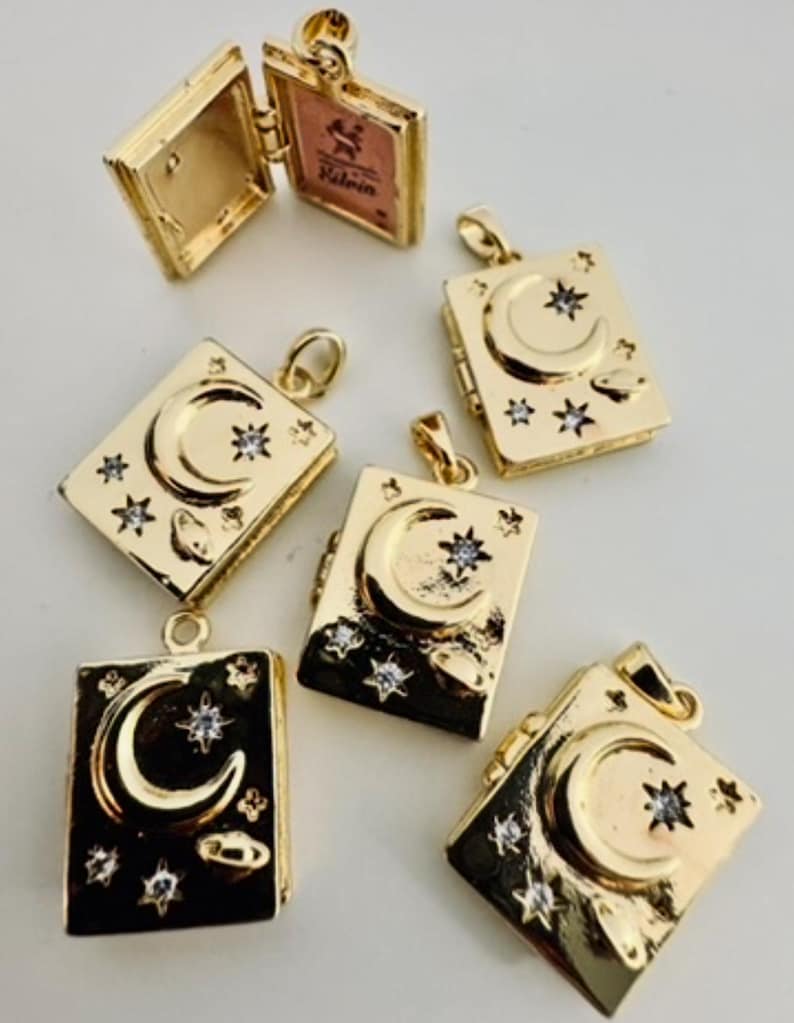 Peut inclure: Pendentifs dor&eacute;s en forme de livre avec un croissant de lune et des &eacute;toiles. Certains pendentifs sont ouverts, r&eacute;v&eacute;lant une petite image. Les pendentifs ont un th&egrave;me c&eacute;leste et sont probablement des bijoux.