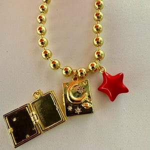Peut inclure: Collier dor&eacute; avec un m&eacute;daillon et des breloques. Le m&eacute;daillon est ouvert, r&eacute;v&eacute;lant un int&eacute;rieur vierge. Un charm rectangulaire repr&eacute;sente un croissant de lune et des &eacute;toiles. Un charm en forme d'&eacute;toile rouge est suspendu &agrave; c&ocirc;t&eacute;.
