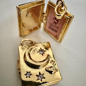 Peut inclure: Pendentif dor&eacute; en forme de livre avec une ouverture &agrave; charni&egrave;re. Un pendentif est ouvert, r&eacute;v&eacute;lant un int&eacute;rieur rose. L'autre pendentif pr&eacute;sente un croissant de lune, des &eacute;toiles et un petit design de plan&egrave;te. Les deux pendentifs ont une boucle pour une cha&icirc;ne.