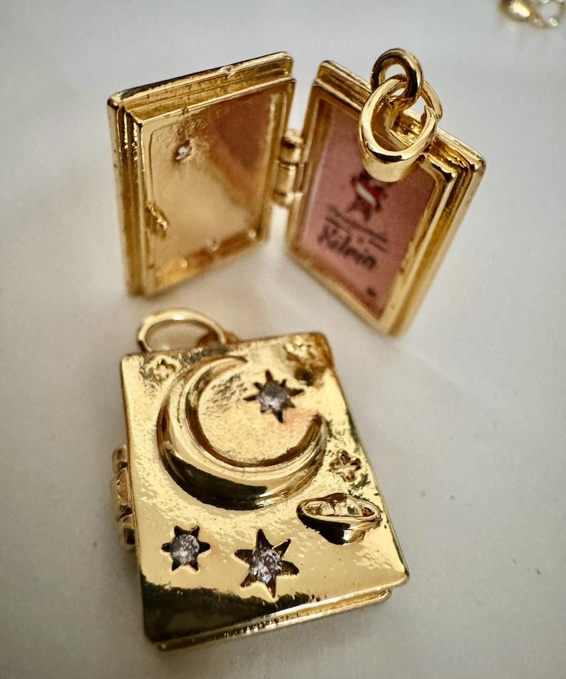 Peut inclure: Pendentif dor&eacute; en forme de livre avec une ouverture &agrave; charni&egrave;re. Un pendentif est ouvert, r&eacute;v&eacute;lant un int&eacute;rieur rose. L'autre pendentif pr&eacute;sente un croissant de lune, des &eacute;toiles et un petit design de plan&egrave;te. Les deux pendentifs ont une boucle pour une cha&icirc;ne.